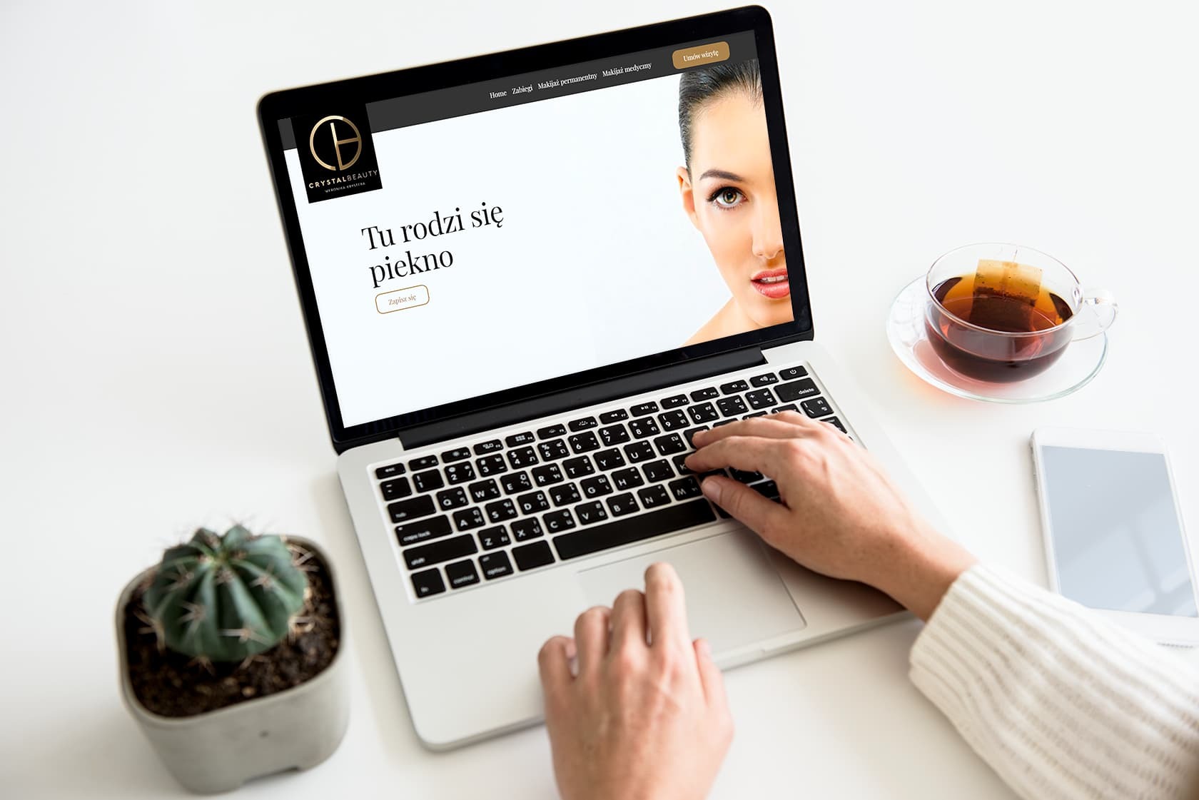 Osoba pisze na laptopie, na ekranie strona internetowa salonu kosmetycznego Crystal Beauty z hasłem 'Tu rodzi się piękno', obok filiżanka herbaty i telefon.