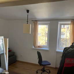 Paweł Paziak - Pokój z dwoma oknami z beżowymi zasłonami, drewnianą podłogą, biurkiem z krzesłem obrotowym i wieszakiem na ubrania, widziany zza framugi drzwi, z widocznym fragmentem sufitu z lampą.