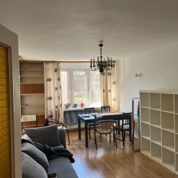 Paweł Paziak - Widok na wnętrze pokoju dziennego z kanapą, stołem jadalnym, regałem i parkietem, oświetlonego naturalnym światłem wpadającym przez okno.