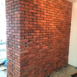 Paweł Paziak - Ściana wykończona czerwoną cegłą klinkierową w narożniku pomieszczenia, widoczne gniazdka elektryczne, folia ochronna na podłodze.