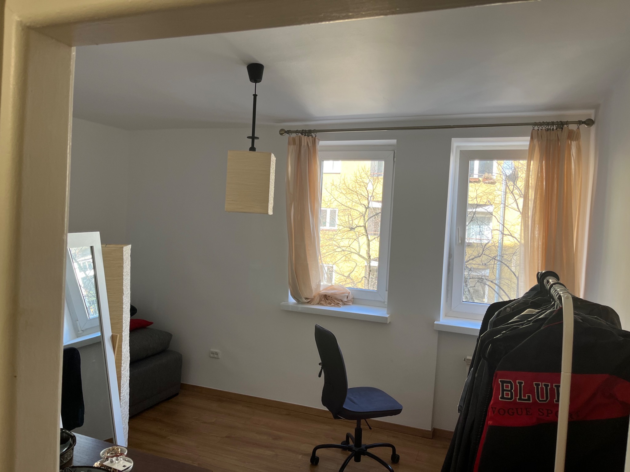 Pokój z dwoma oknami z beżowymi zasłonami, drewnianą podłogą, biurkiem z krzesłem obrotowym i wieszakiem na ubrania, widziany zza framugi drzwi, z widocznym fragmentem sufitu z lampą.