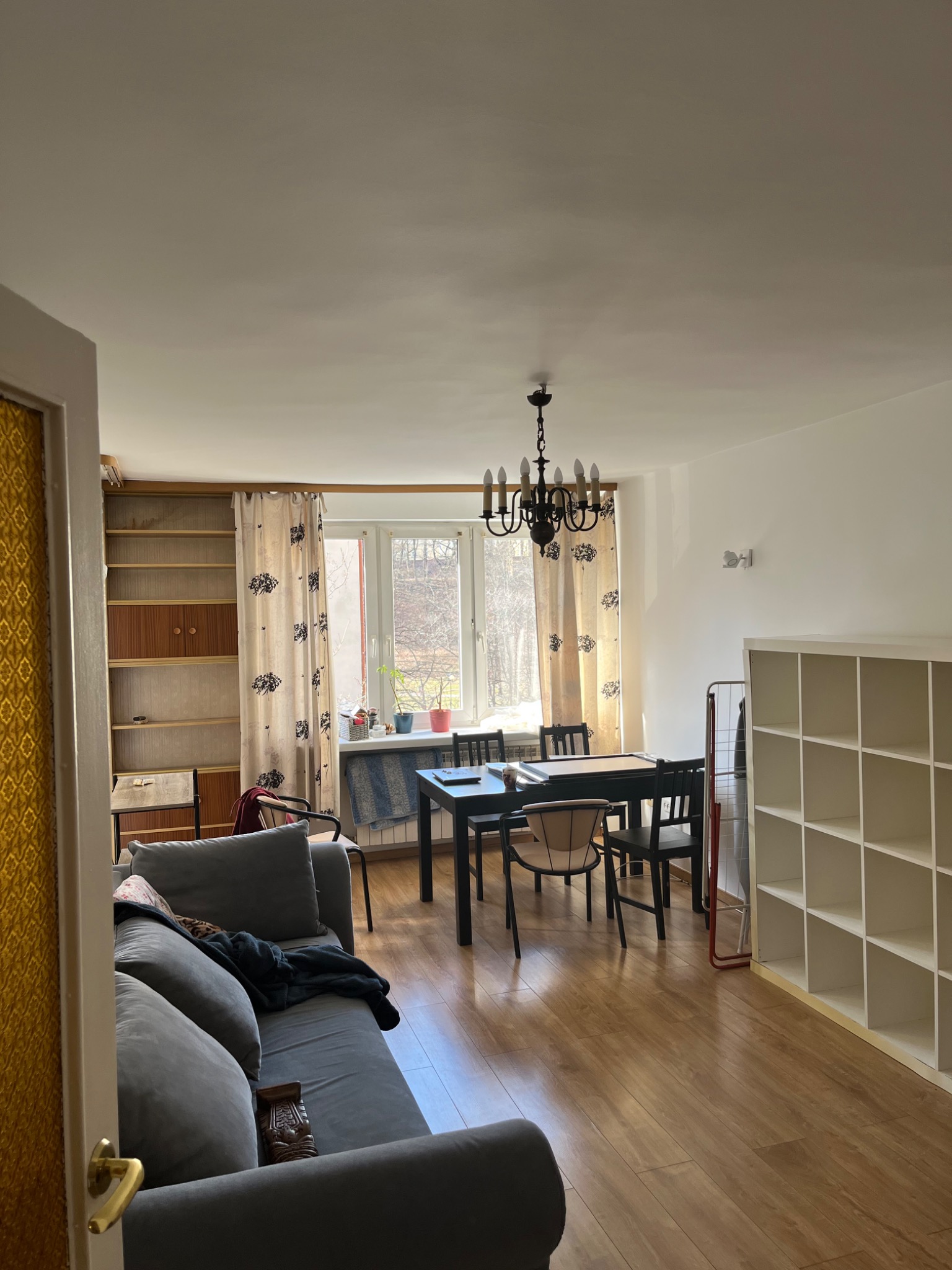 Widok na wnętrze pokoju dziennego z kanapą, stołem jadalnym, regałem i parkietem, oświetlonego naturalnym światłem wpadającym przez okno.