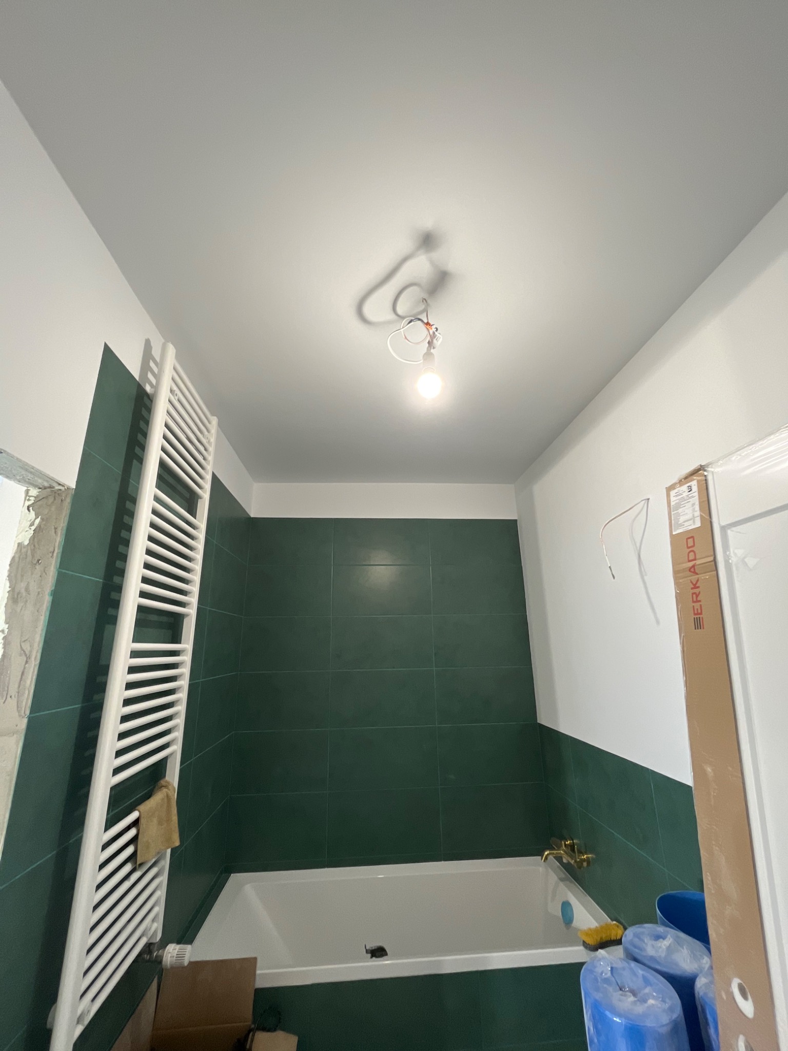 Wnętrze łazienki w trakcie remontu: zielone płytki do połowy ściany, biała wanna, grzejnik drabinkowy, wisząca żarówka bez klosza, kartonowe pudła i niebieskie wiadra na podłodze.