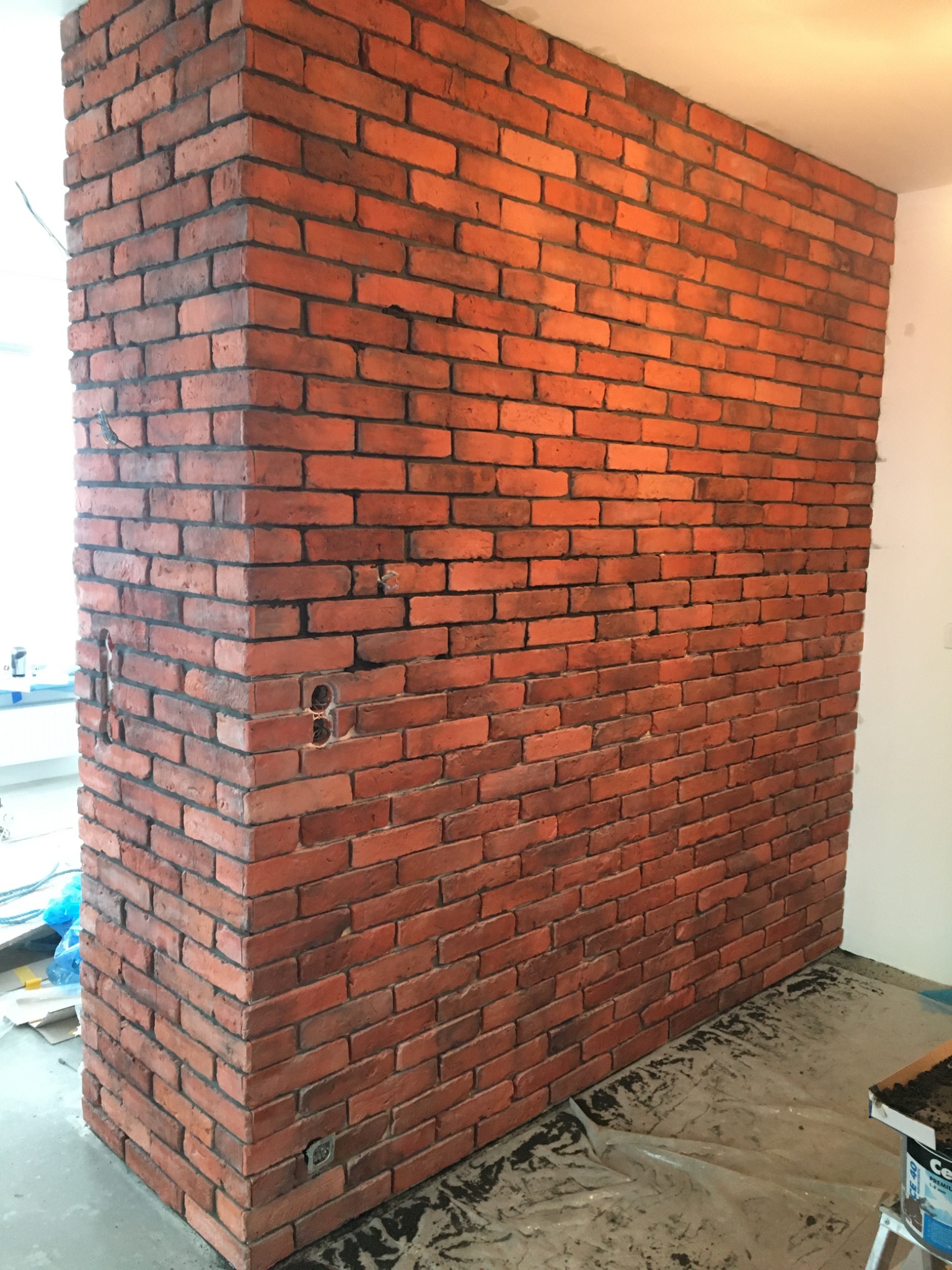 Ściana wykończona czerwoną cegłą klinkierową w narożniku pomieszczenia, widoczne gniazdka elektryczne, folia ochronna na podłodze.