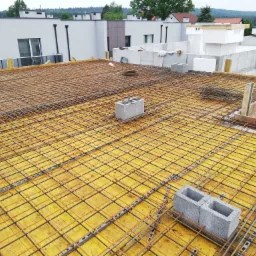 Zbrojenie stropu domu jednorodzinnego z widoczną siatką z prętów stalowych, deskowaniem z żółtych płyt, bloczkami betonowymi i elementami szalunkowymi, w tle budynki mieszkalne.