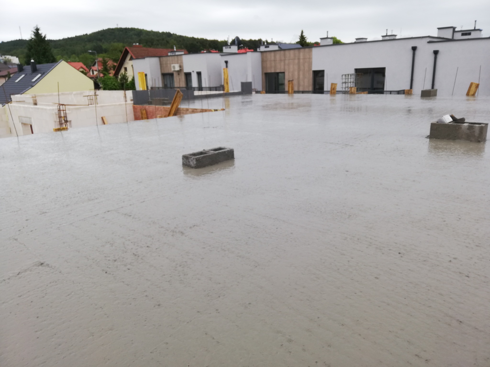 Świeżo wylany betonowy strop na budowie domu z widocznymi bloczkami betonowymi, w tle zabudowa jednorodzinna i zalesione wzgórze, pochmurny dzień.