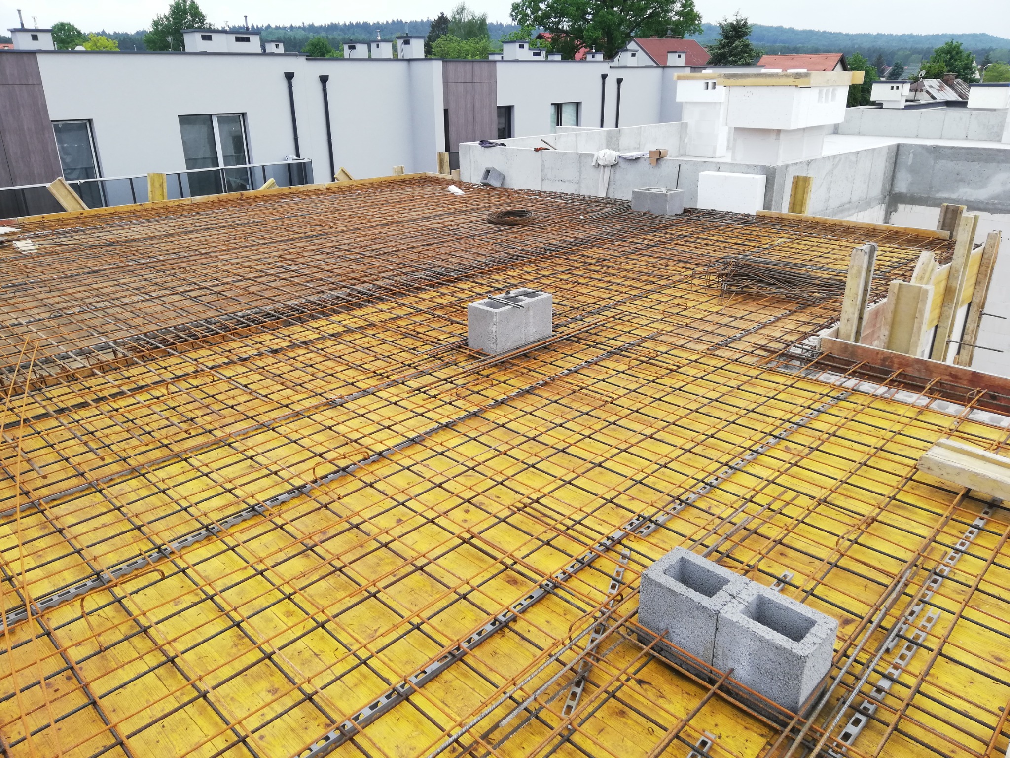 Zbrojenie stropu domu jednorodzinnego z widoczną siatką z prętów stalowych, deskowaniem z żółtych płyt, bloczkami betonowymi i elementami szalunkowymi, w tle budynki mieszkalne.