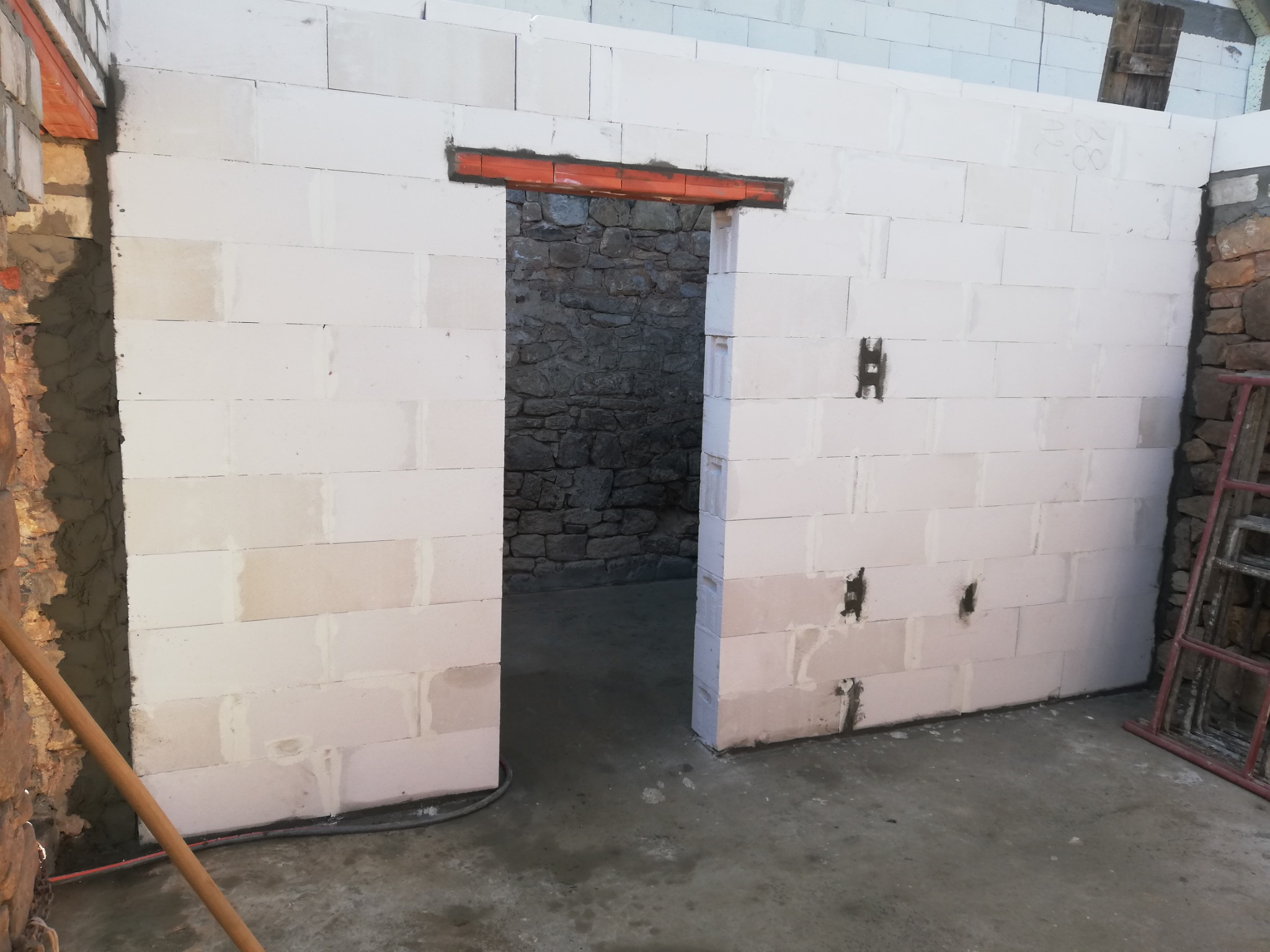 Wznoszenie ściany z białych bloczków, widoczne nadproże drzwiowe z cegieł, surowe wnętrze z kamiennymi ścianami w tle, drabina oparta o ścianę.