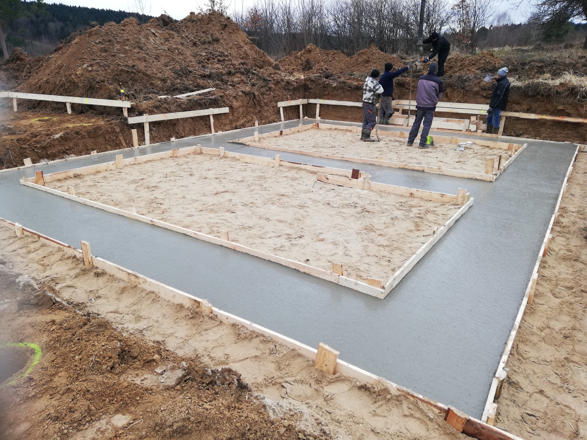 Wylany fundament pod budynek z drewnianym szalunkiem i pracownikami budowlanymi na placu budowy, widoczny duży nasyp ziemi w tle.