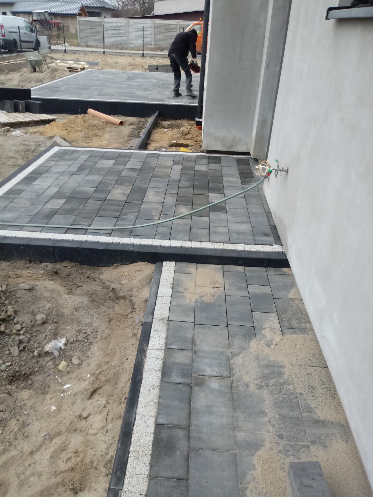 Układanie szarej kostki brukowej z białymi obrzeżami przy ścianie budynku, widoczny pracownik w trakcie pracy i przygotowany teren pod dalsze prace.