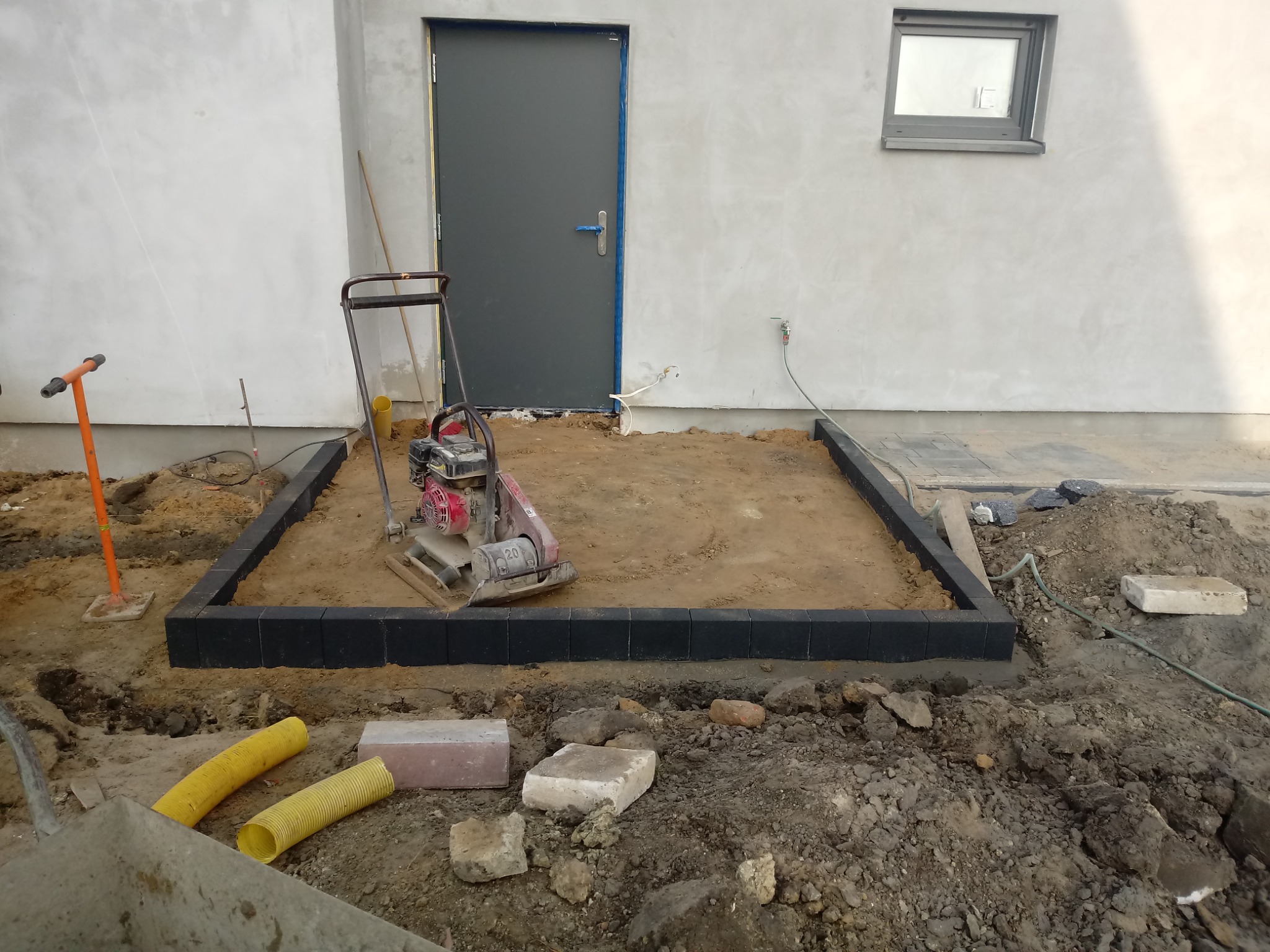Przygotowanie podłoża pod taras: ubijarka wibracyjna na piasku, obrzeża z ciemnych kostek, żółte rury drenażowe i narzędzia budowlane obok ściany budynku z szarą stolarką okienną i drzwiową.