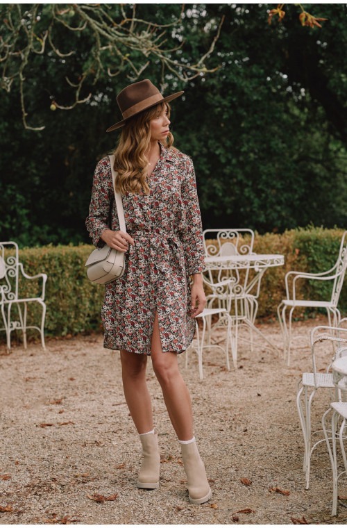 Kobieta we Wrocławiu pozuje w kwiecistej sukience koszulowej z paskiem, brązowym kapeluszu i beżowych botkach, z kremową torebką na ramieniu, na tle ogrodu z białymi metalowymi meblami.