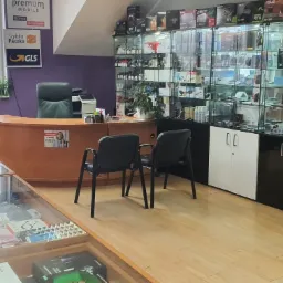 Wnętrze sklepu komputerowego z ladą obsługi klienta, gablotami pełnymi podzespołów komputerowych (kart graficznych, procesorów) oraz oznakowaniem usług kurierskich na ścianie.