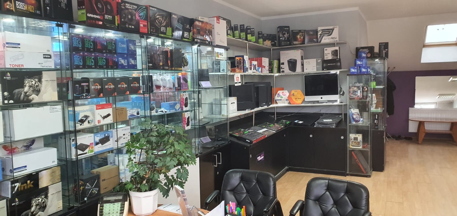 Wnętrze sklepu komputerowego z przeszklonymi gablotami i półkami, na których eksponowane są procesory Intel Core i Ryzen, karty graficzne RTX, tonery do drukarek i inne komponenty komputerowe.