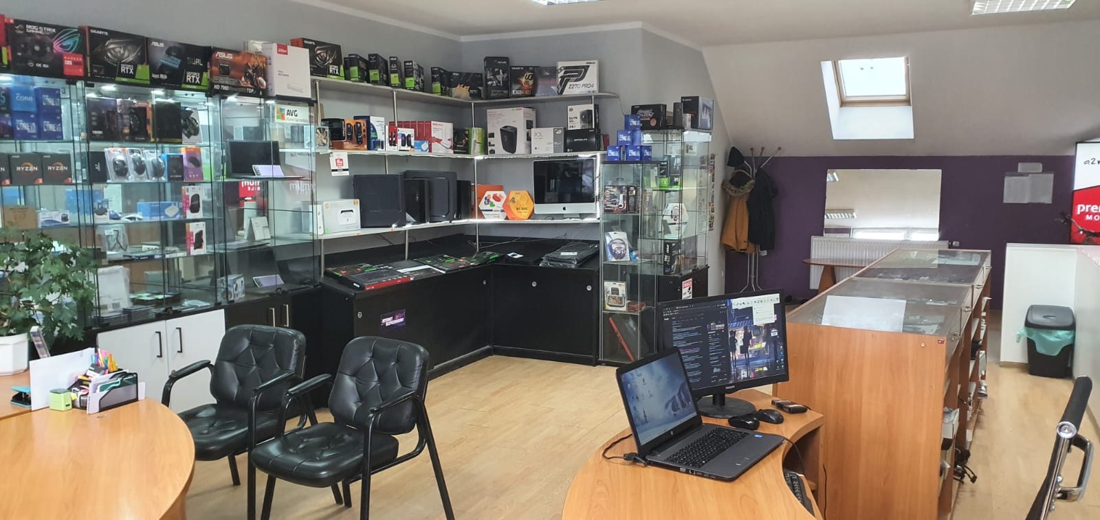 Wnętrze sklepu komputerowego z ekspozycją komponentów, laptopem i monitorem na pierwszym planie oraz regałami pełnymi pudełek po podzespołach w tle.