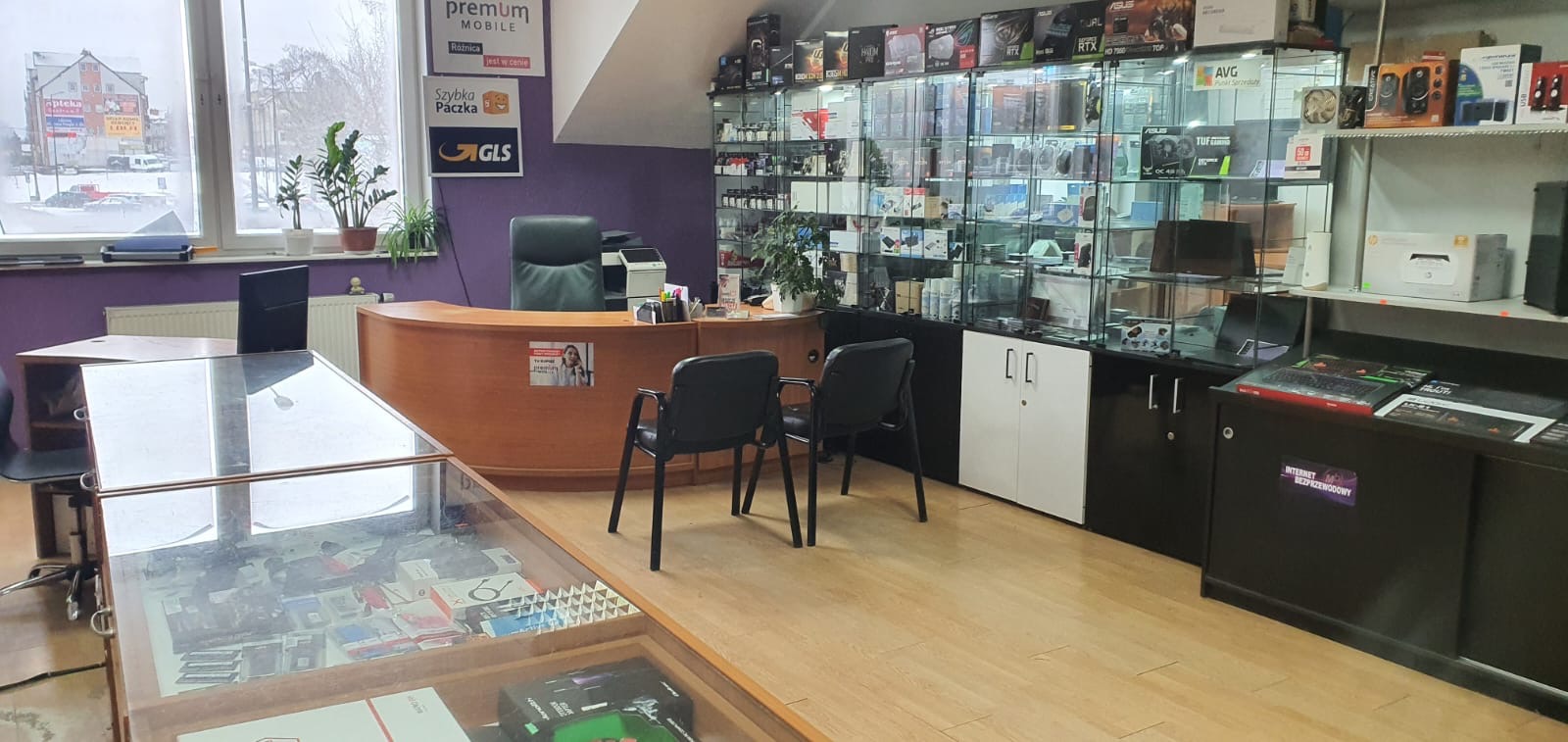 Wnętrze sklepu komputerowego z ladą obsługi klienta, gablotami pełnymi podzespołów komputerowych (kart graficznych, procesorów) oraz oznakowaniem usług kurierskich na ścianie.