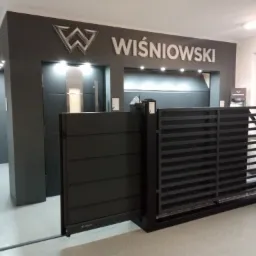 Ekspozycja dwóch modeli bram: pełnej, poziomej i ażurowej, w salonie firmowym z logo Wiśniowski na ścianie.