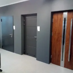 Ekspozycja trzech różnych modeli drzwi w salonie: ciemnoszare drzwi gładkie, szare drzwi panelowe i drzwi drewniane z metalowymi wstawkami.