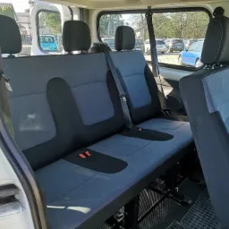 Wnętrze busa z trzema rzędami szarych siedzeń, widok z otwartych drzwi bocznych na zaparkowane samochody w tle.