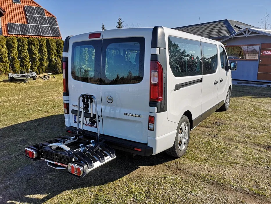 Biały bus Opel Vivaro z zamontowanym bagażnikiem rowerowym Thule na haku holowniczym, stojący na trawniku przed domem z panelami słonecznymi na dachu.