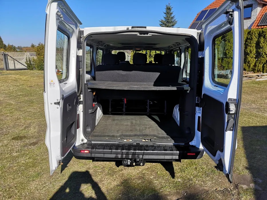 Wnętrze białego busa z otwartymi drzwiami, widok z tyłu na puste siedzenia i przestrzeń ładunkową, hak holowniczy na zewnątrz, zielony trawnik i dom z panelami słonecznymi w tle.