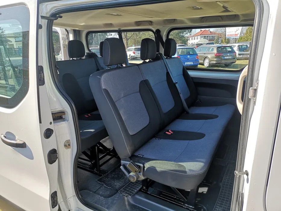 Wnętrze białego busa z trzema rzędami siedzeń w odcieniach szarości i czerni, widok z otwartych drzwi bocznych na zaparkowane samochody i budynki w tle.