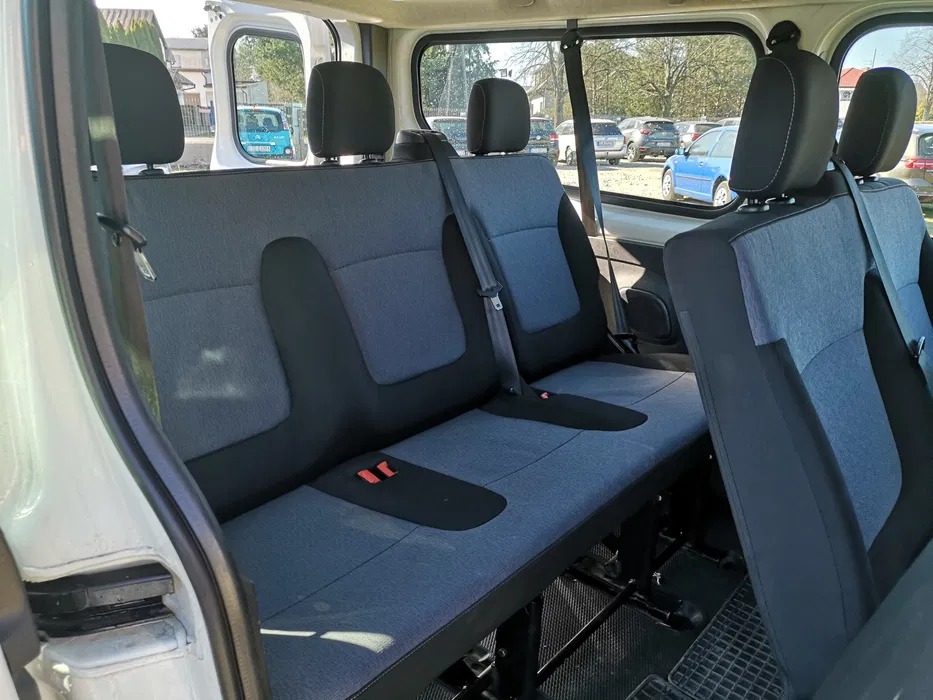 Wnętrze busa z trzema rzędami szarych siedzeń, widok z otwartych drzwi bocznych na zaparkowane samochody w tle.