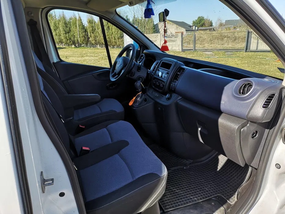 Wnętrze białego busa z niebieskimi siedzeniami i czarną deską rozdzielczą, widok z otwartych drzwi kierowcy, na lusterku wisi breloczek.