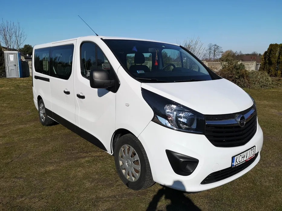 Biały Opel Vivaro, bus do przewozu osób, zaparkowany na trawie w słoneczny dzień, widoczna przenośna toaleta w tle.