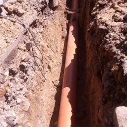 Pomarańczowa rura kanalizacyjna ułożona w wąskim wykopie w ziemi, widoczne fragmenty betonu i gruzu po bokach wykopu.