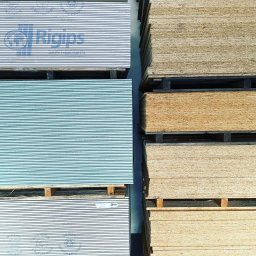 Solid Ostrów Materiały Budowlane - Stosy płyt gipsowo-kartonowych marki Rigips na paletach, ułożone obok stosu płyt drewnopochodnych.