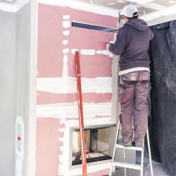 Solid Ostrów Materiały Budowlane - Pracownik na drabinie rysuje linię na płycie kartonowo-gipsowej przy kominku, widoczny poziomica, szpachlowane łączenia płyt.