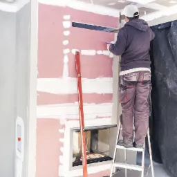 Pracownik na drabinie rysuje linię na płycie kartonowo-gipsowej przy kominku, widoczny poziomica, szpachlowane łączenia płyt.