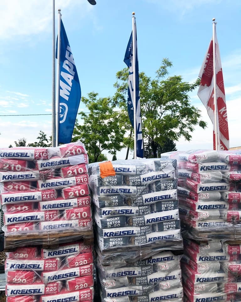 Sztaple worków z zaprawą i betonem marki Kreisel, ułożone na paletach przed składem budowlanym, z widocznymi flagami reklamowymi producentów i pomarańczowym kaskiem na jednym ze stosów.