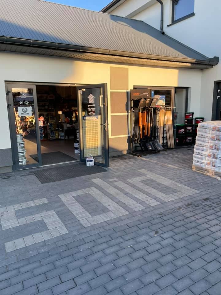 Otwarty sklep budowlany z ekspozycją narzędzi i materiałów, widoczny napis 'SOLID' ułożony z kostki brukowej przed wejściem.