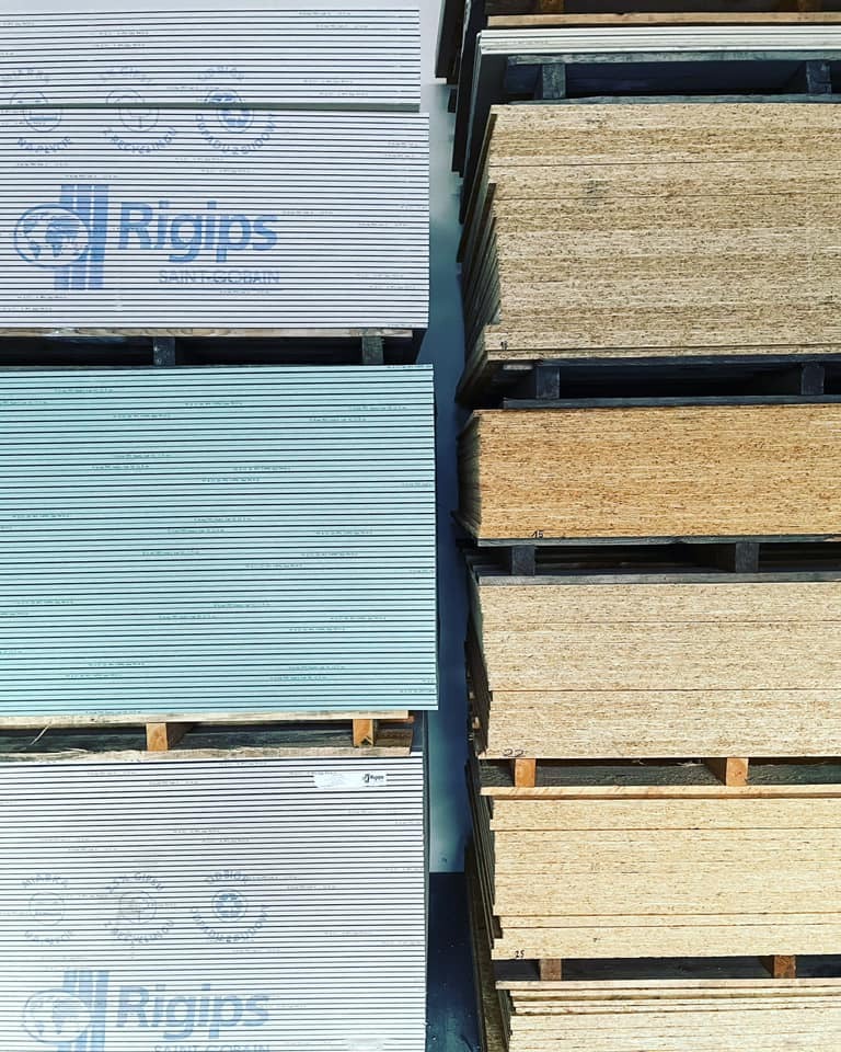 Stosy płyt gipsowo-kartonowych marki Rigips na paletach, ułożone obok stosu płyt drewnopochodnych.