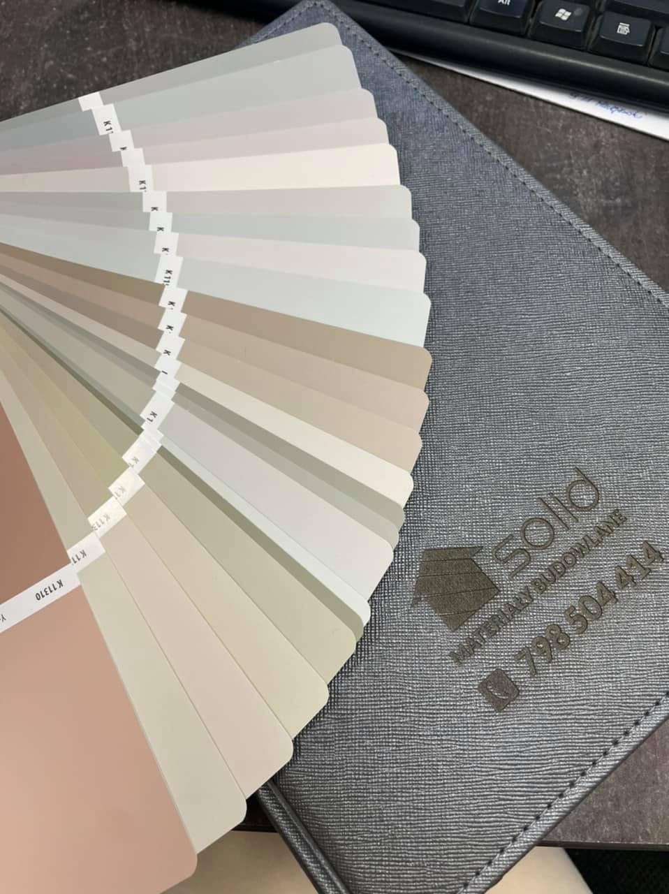 Paleta próbek kolorów farb w odcieniach beżu, szarości i różu, ułożona wachlarzowo na szarym etui z logo firmy 'Solid Materiały Budowlane' i numerem telefonu.