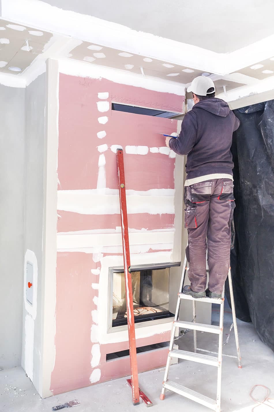 Pracownik na drabinie rysuje linię na płycie kartonowo-gipsowej przy kominku, widoczny poziomica, szpachlowane łączenia płyt.