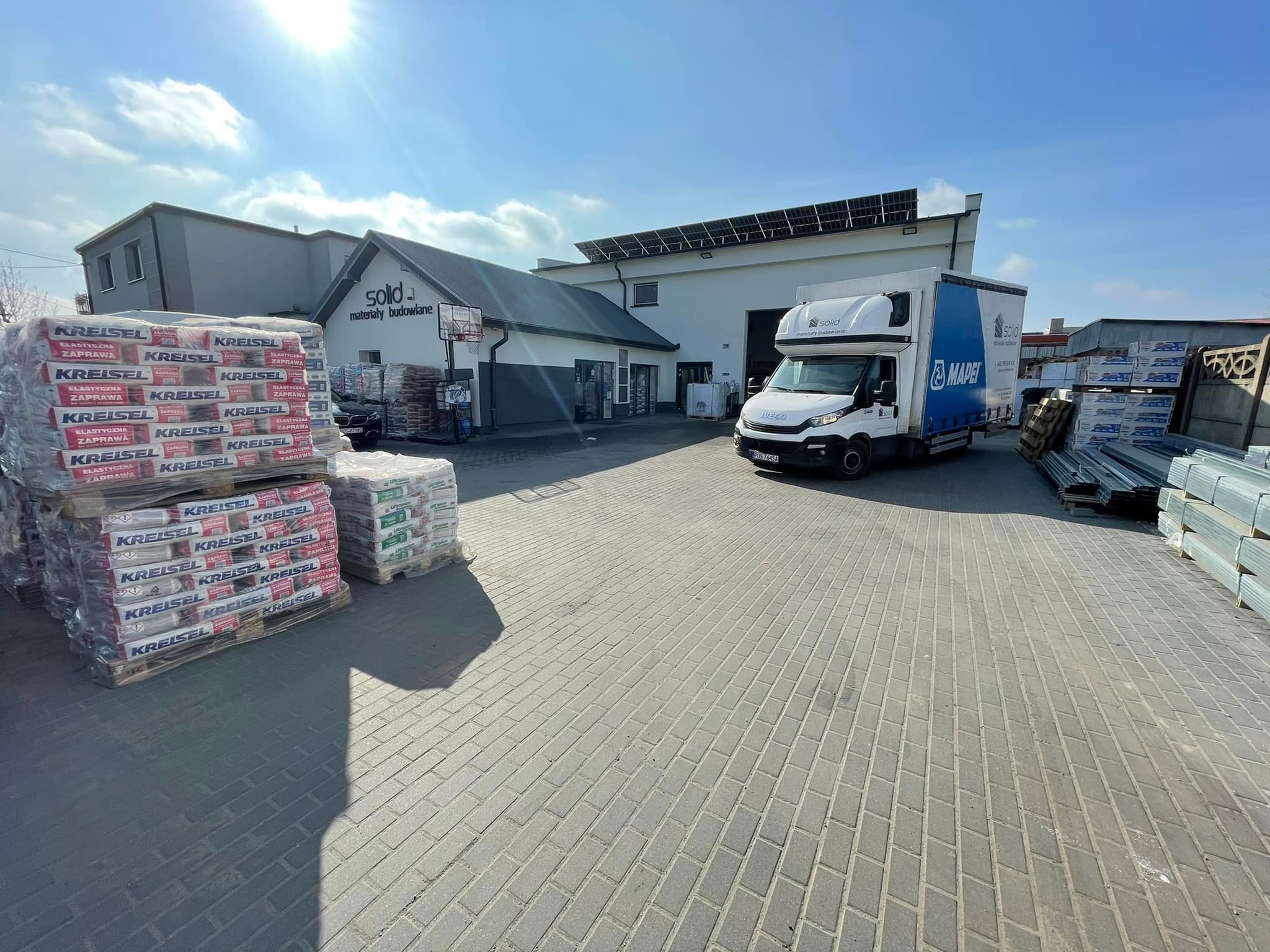 Skład budowlany z zapasem zapraw marki Kreisel na paletach, biała ciężarówka dostawcza z logo Mapei w tle, budynek z panelami słonecznymi na dachu.