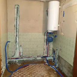 Wnętrze łazienki w trakcie remontu z widoczną instalacją wodną i grzewczą: biały bojler, niebieski zbiornik wyrównawczy, rury w izolacji piankowej, bruzdy w ścianach wypełnione pianą montażową...