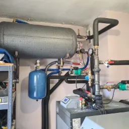 Złożona instalacja grzewcza z szarym zbiornikiem akumulacyjnym, niebieskim naczyniem przeponowym i piecem w kotłowni, widoczne liczne rury, zawory i pompy obiegowe.