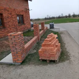 Wznoszenie muru z czerwonej cegły przy domu jednorodzinnym, widoczne słupki i fragment muru, paleta z cegłami obok, w tle krajobraz wiejski.