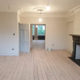 Jasne, przestronne wnętrze po odświeżeniu, z pomalowanymi na biało ścianami, drewnianą podłogą, ozdobnym kominkiem, sztukaterią sufitową i otwartymi drzwiami do kolejnego pomieszczenia. Widoczna...