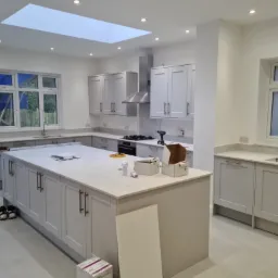 Jasna kuchnia w trakcie montażu, z wyspą, szarymi szafkami i oknem dachowym, widoczne narzędzia i materiały montażowe.