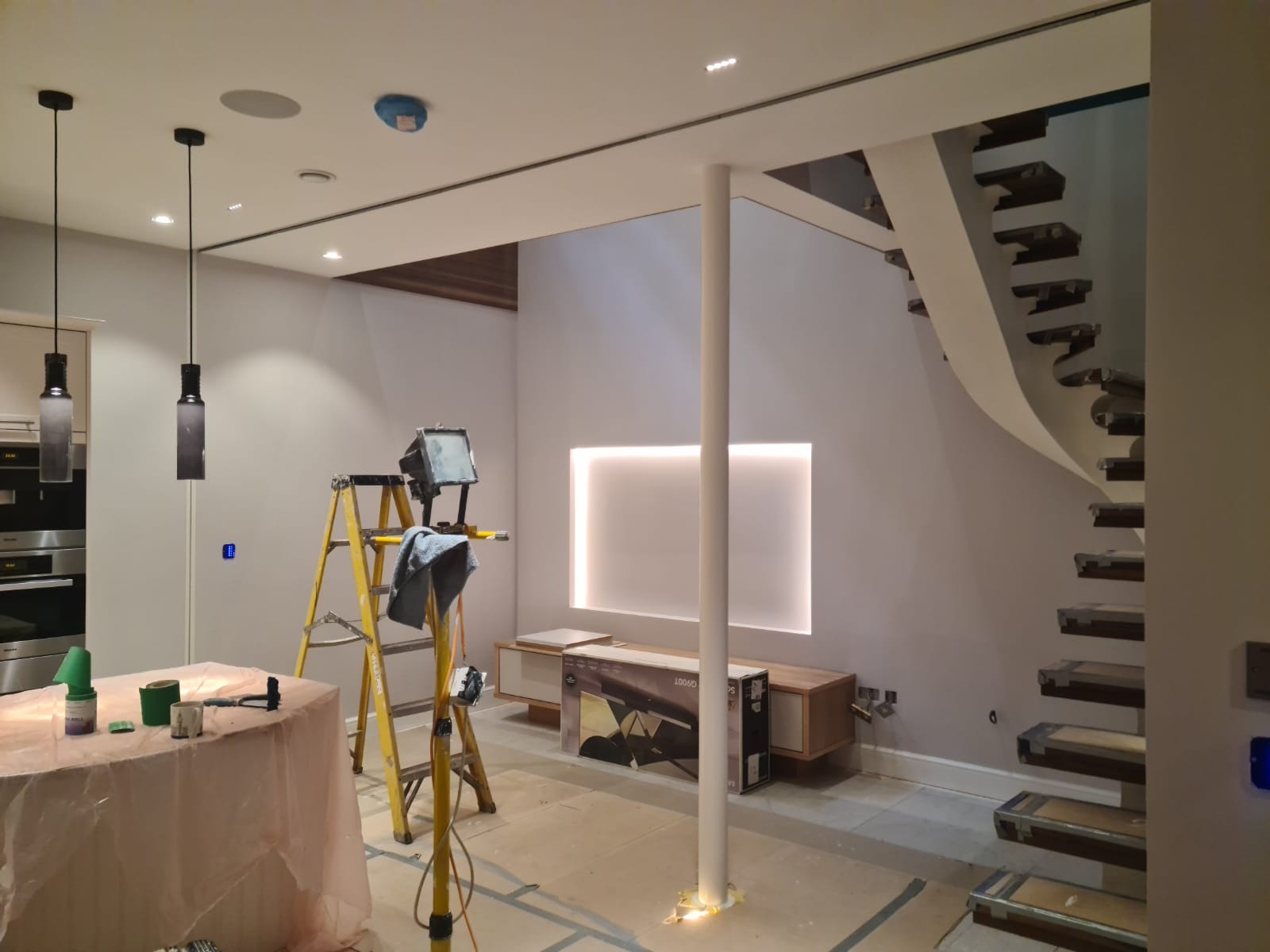 Wnętrze w trakcie prac wykończeniowych: zabezpieczone meble, drabina z lampą, widoczne materiały malarskie, nowoczesne schody spiralne i podświetlona wnęka w ścianie.
