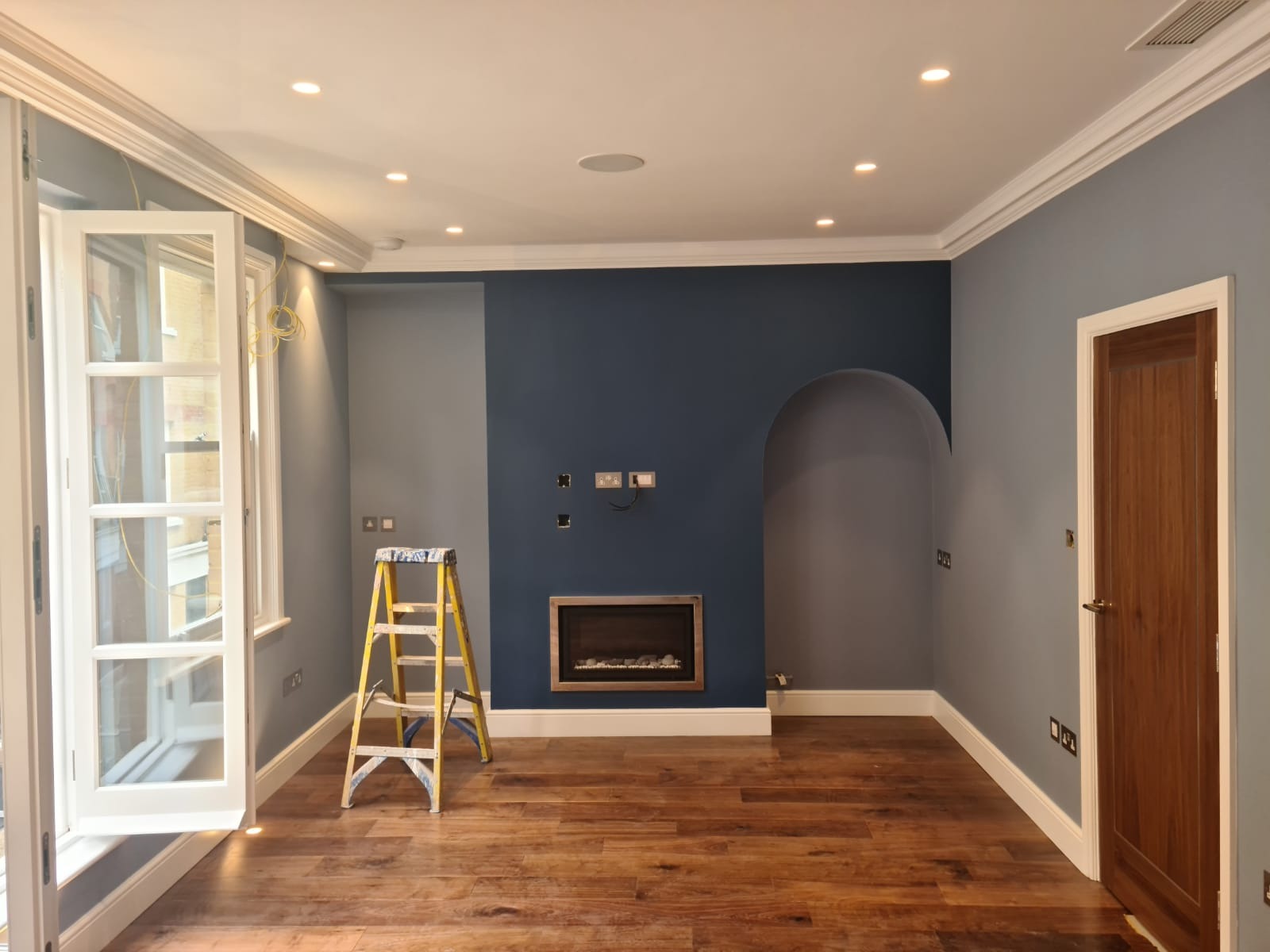 Pokój po remoncie: świeżo pomalowane ściany w odcieniach szarości i granatu, drewniana podłoga, drabina malarska stoi przy oknie, widoczne gniazdka elektryczne przed montażem.