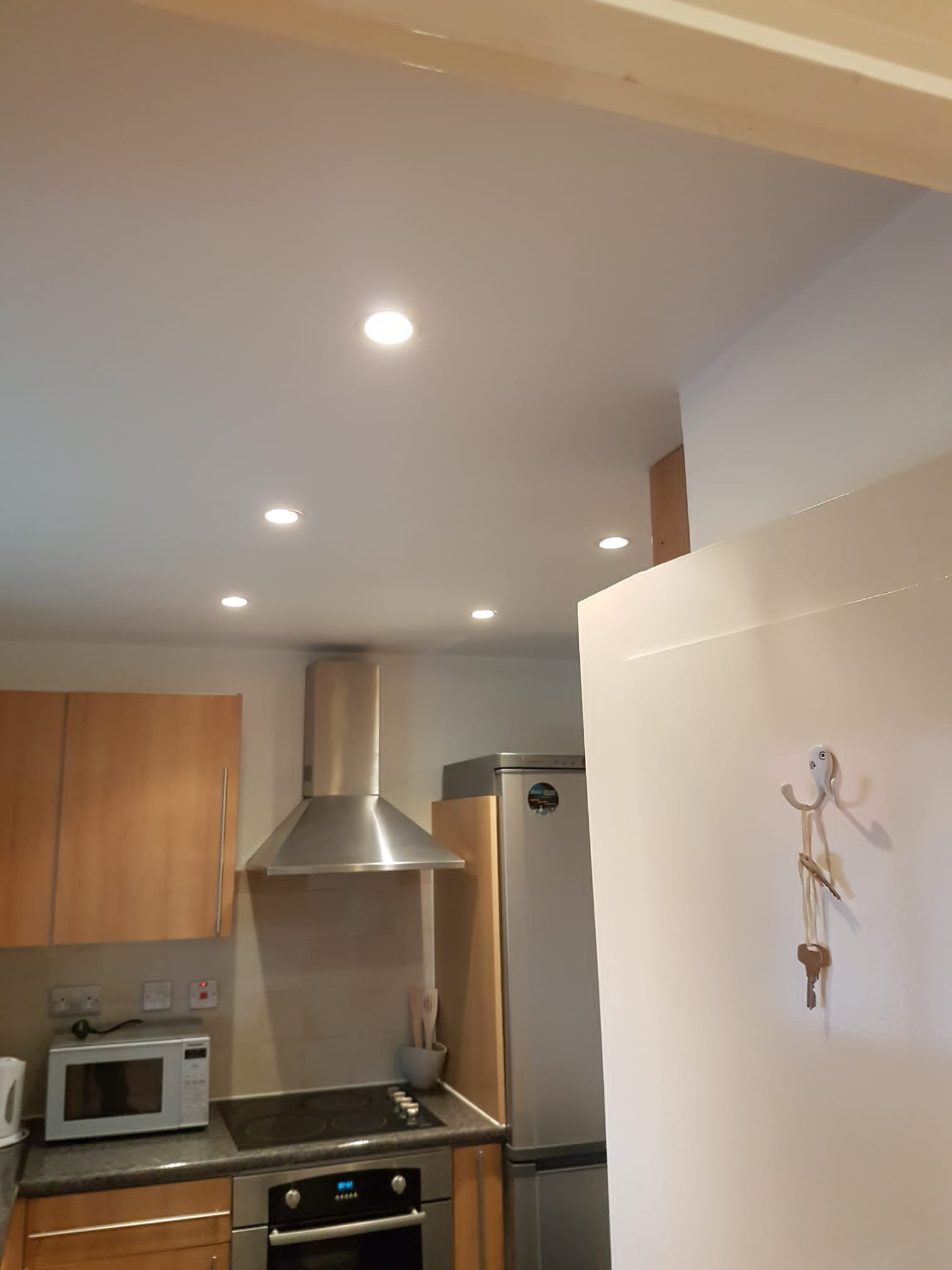 Kuchnia z nowo zamontowanymi punktowymi lampami sufitowymi LED, widoczny fragment szafek kuchennych, okap, kuchenka i lodówka. Na ścianie wisi haczyk z kluczami.
