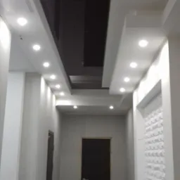 Korytarz w trakcie remontu z czarnym podwieszanym sufitem i białymi ścianami, oświetlony rzędem punktowych lamp halogenowych, widoczne ślady po pracach na podłodze.