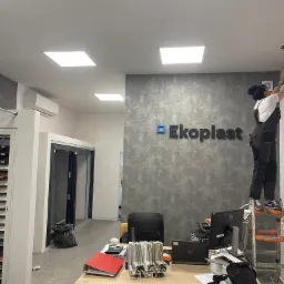 Pracownica na drabinie montuje logo 'Ekoplast' na szarej, teksturowanej ścianie w biurze. Widoczny biurko z komputerem i dokumentami, a w tle przeszklone drzwi.