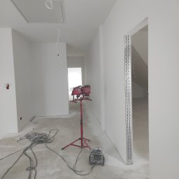 All-Bud Aleksander Sobczak - Wnętrze w trakcie wykańczania: białe ściany po szpachlowaniu i malowaniu, widoczne kable, lampa budowlana na trójnogu, szlifierka do gipsu oraz metalowe profile ościeżnic.